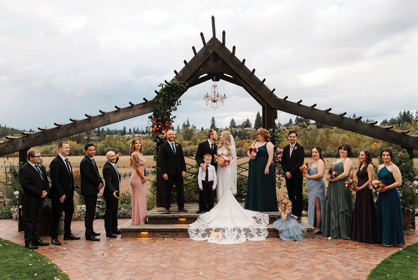 Wedding party poses outdoors in a line at a scenic vineyard venue with mountains in the background during sunset.