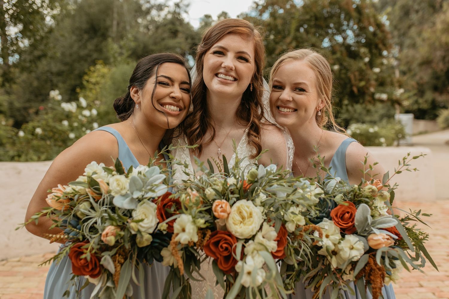 Three bridesmaids in blue dresses smiling and holding lush bouquets with rust, white, and green flowers outdoors.