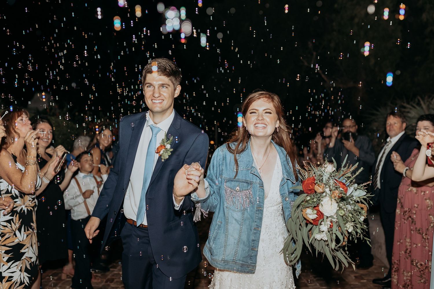 Joyful newlyweds walk through bubbles, bride in denim jacket holding bouquet, groom in navy suit, guests cheering.