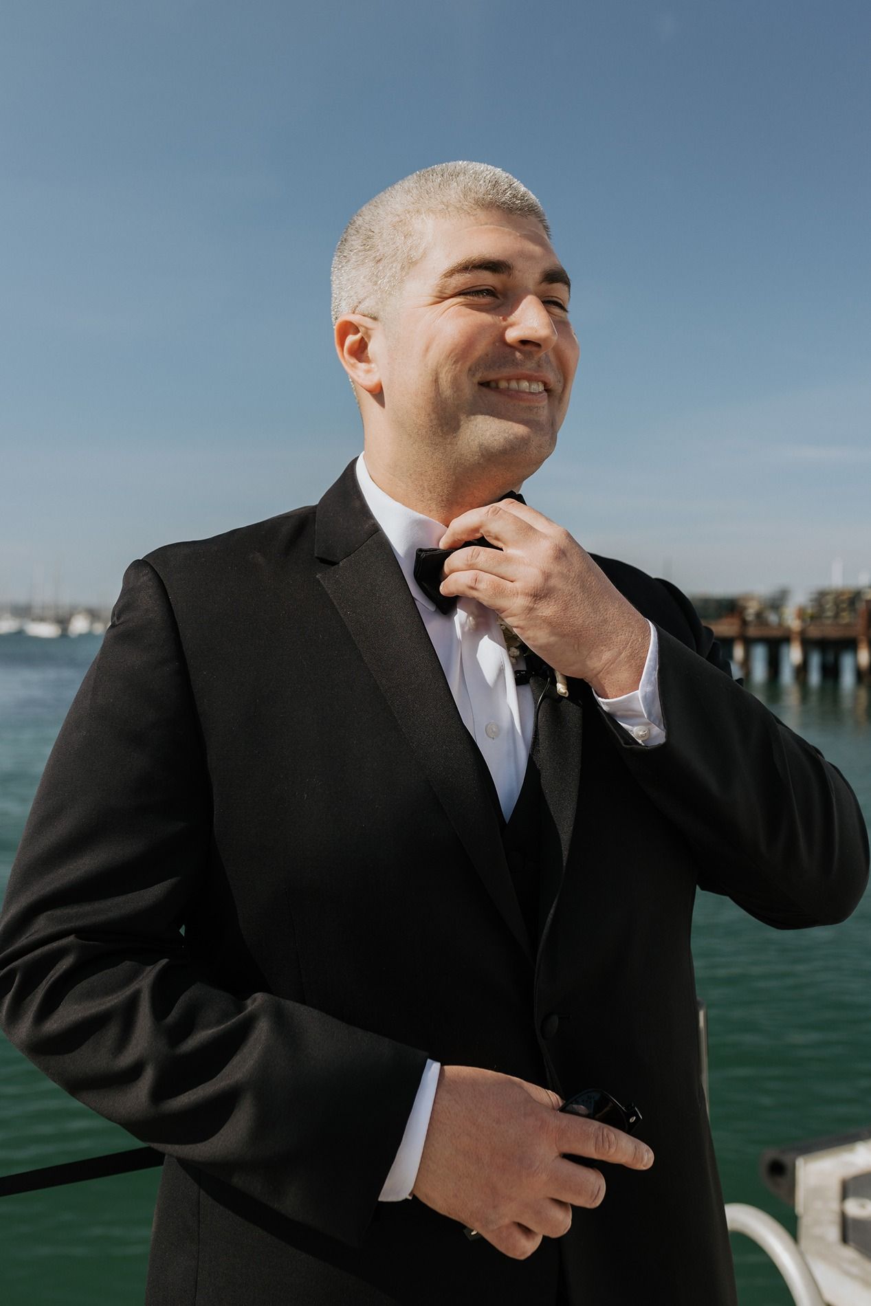 A groom in a black suit adjusts his tie with turquoise water in the background in San Diego.