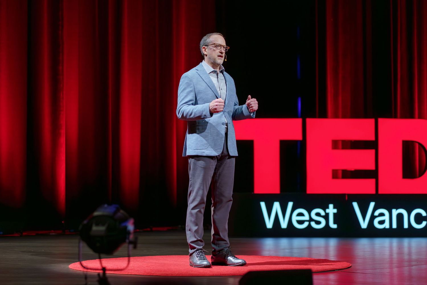 A speaker presents on stage at a TEDx event with a red backdrop and "West Vanc" visible in blue text.