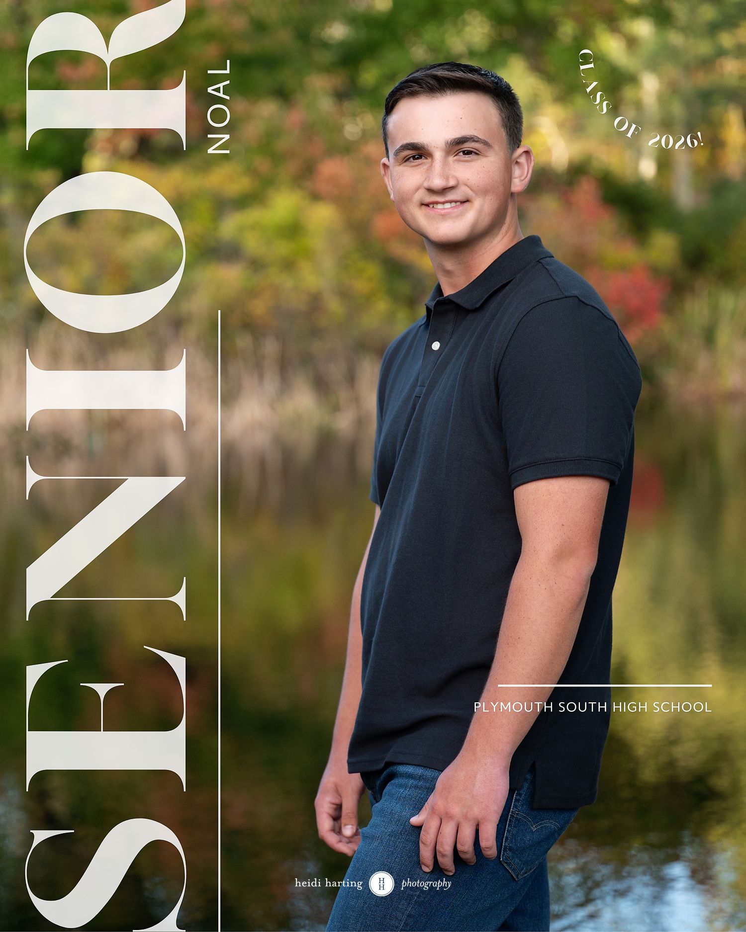 High school senior portrait photo in black polo shirt standing by lake with autumn foliage in background.