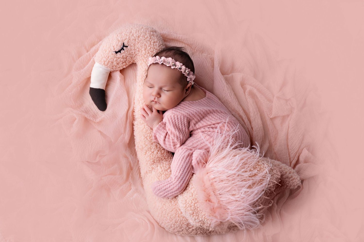 A sleeping newborn baby swaddled in pink fabric lies peacefully on a soft blanket in a studio setting.