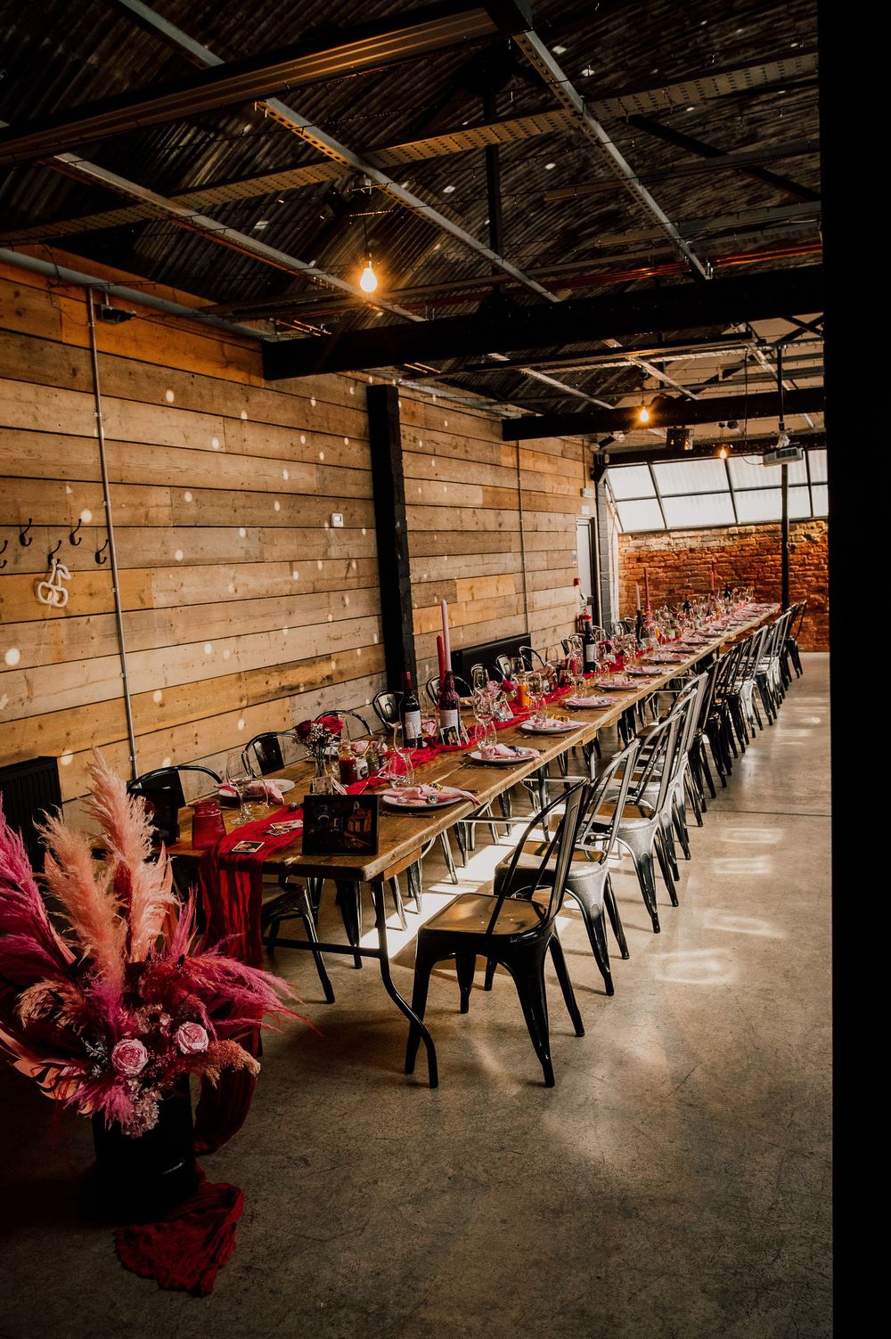 Long rustic reception venue with wooden walls and tables arranged in rows, decorated with red accents and ambient lighting.