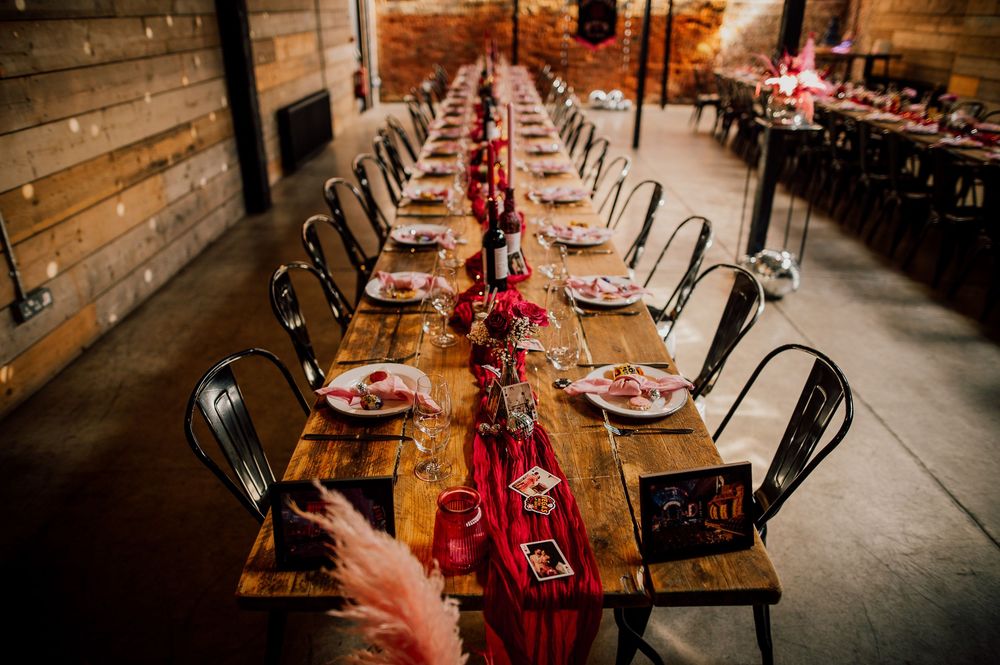 Long wooden dining tables set for an elegant rustic wedding reception with red floral centerpieces and industrial decor.