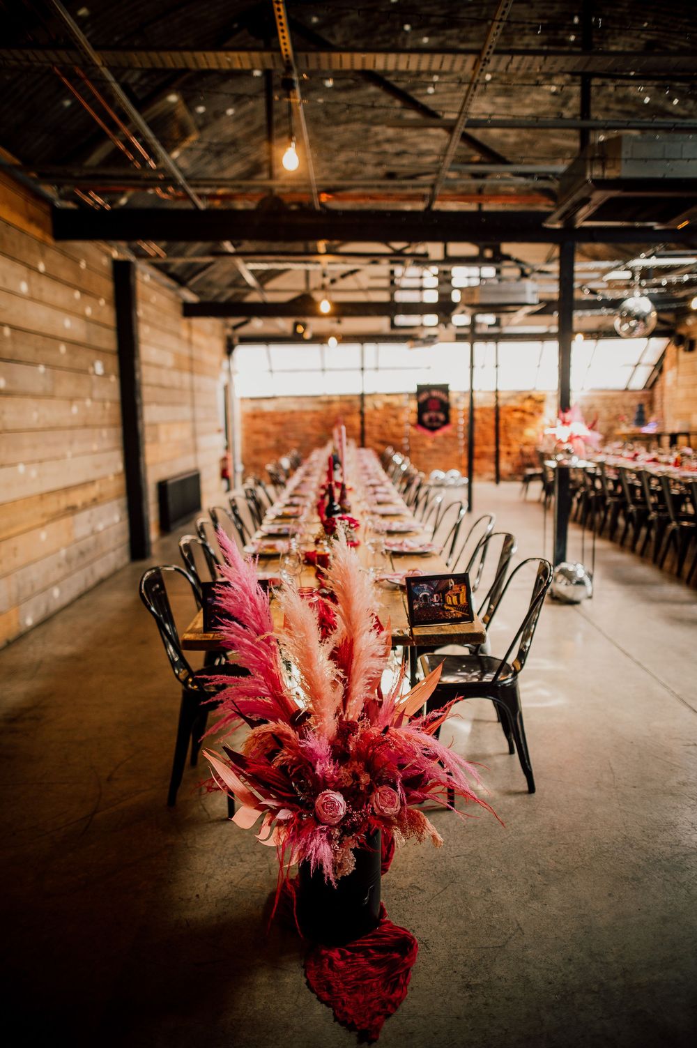 Long rustic wooden tables with black chairs and dramatic red floral centerpieces create an industrial chic event space.