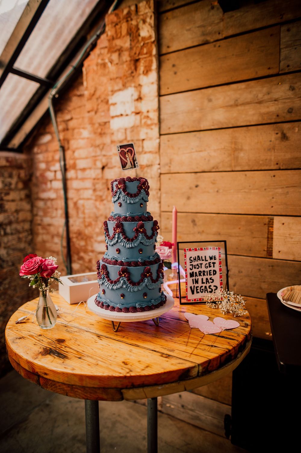 A blue tiered wedding cake displayed on a rustic wooden table with brick wall backdrop.