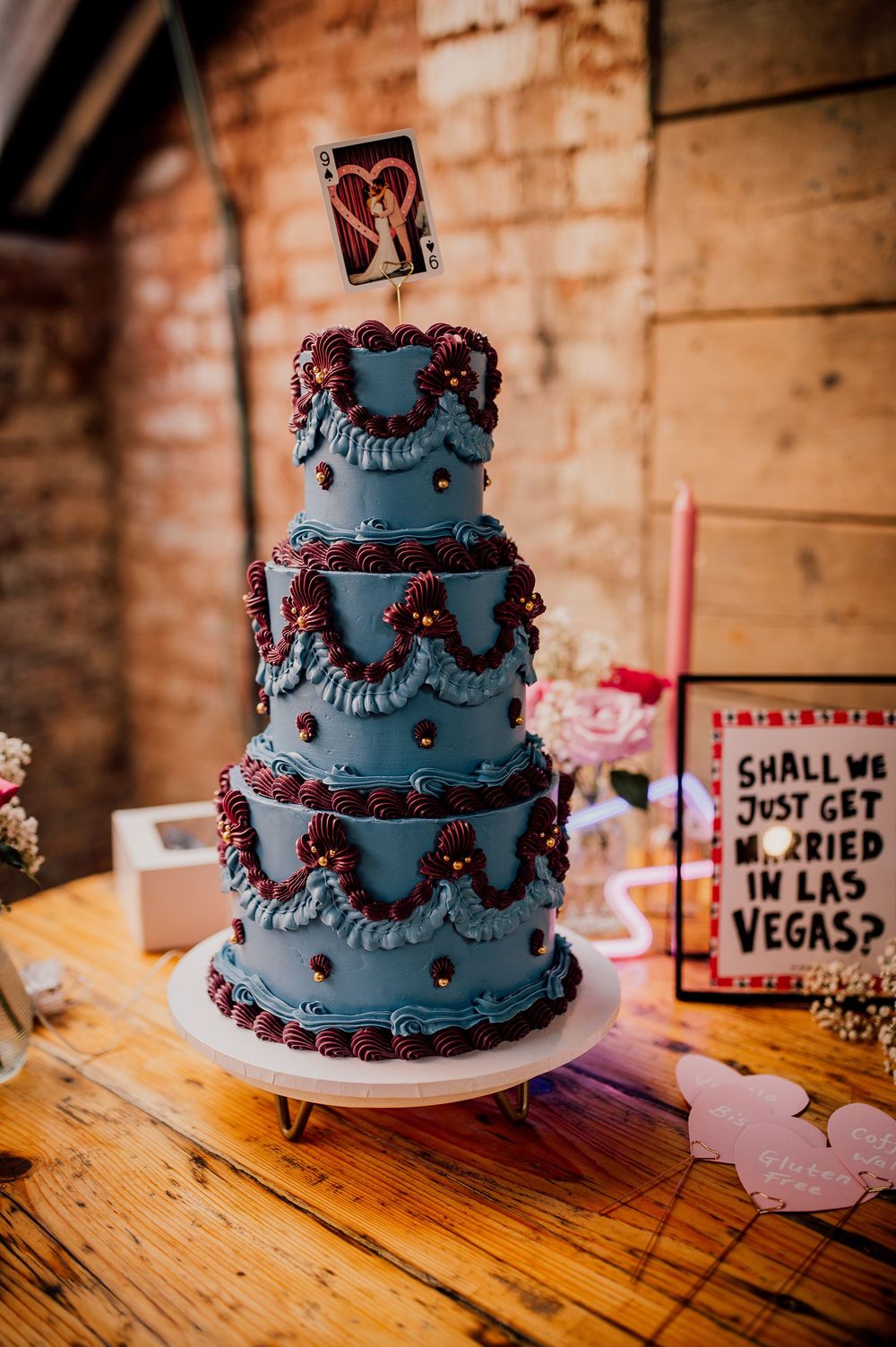 A three-tiered black wedding cake with red accents sits on a wooden table with Las Vegas themed decor nearby.