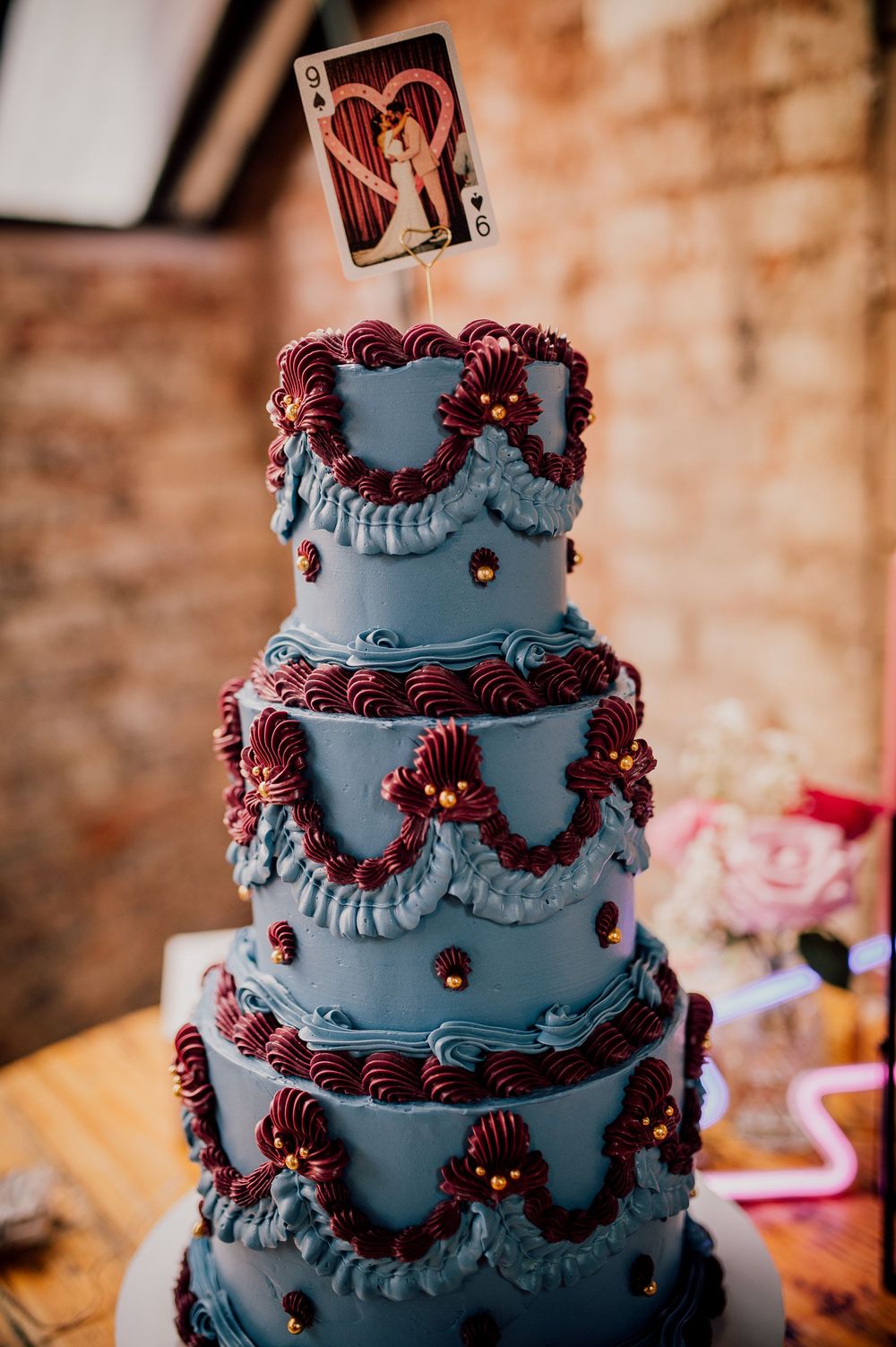 A dark blue three-tiered wedding cake decorated with burgundy piping and floral accents sits on a rustic wooden table.