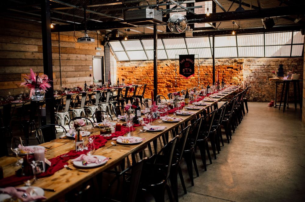 Long rustic wooden tables with pink place settings line a dimly lit industrial-style venue with exposed brick walls.