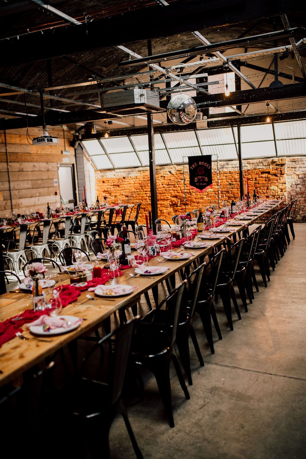 Long rustic dining tables with black chairs and pink table runners set up in an industrial warehouse venue for an event.