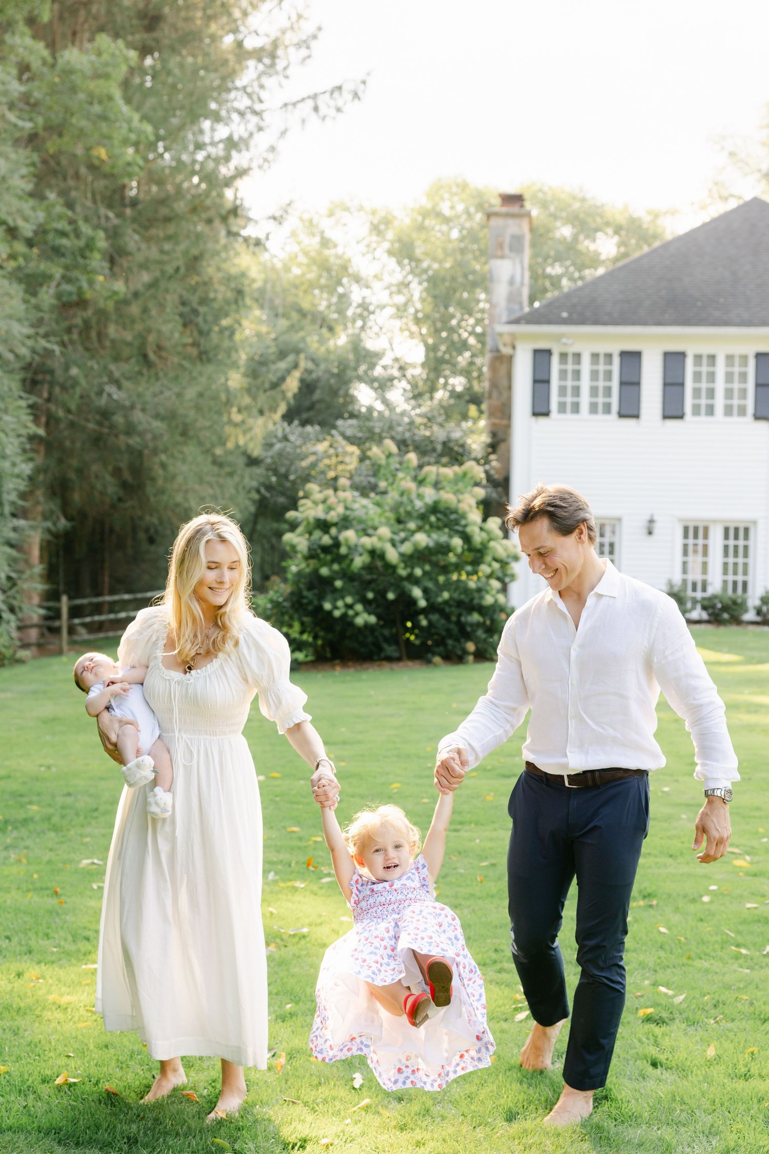 A family in white and navy outfits plays together on a sunny lawn outside a traditional white house.