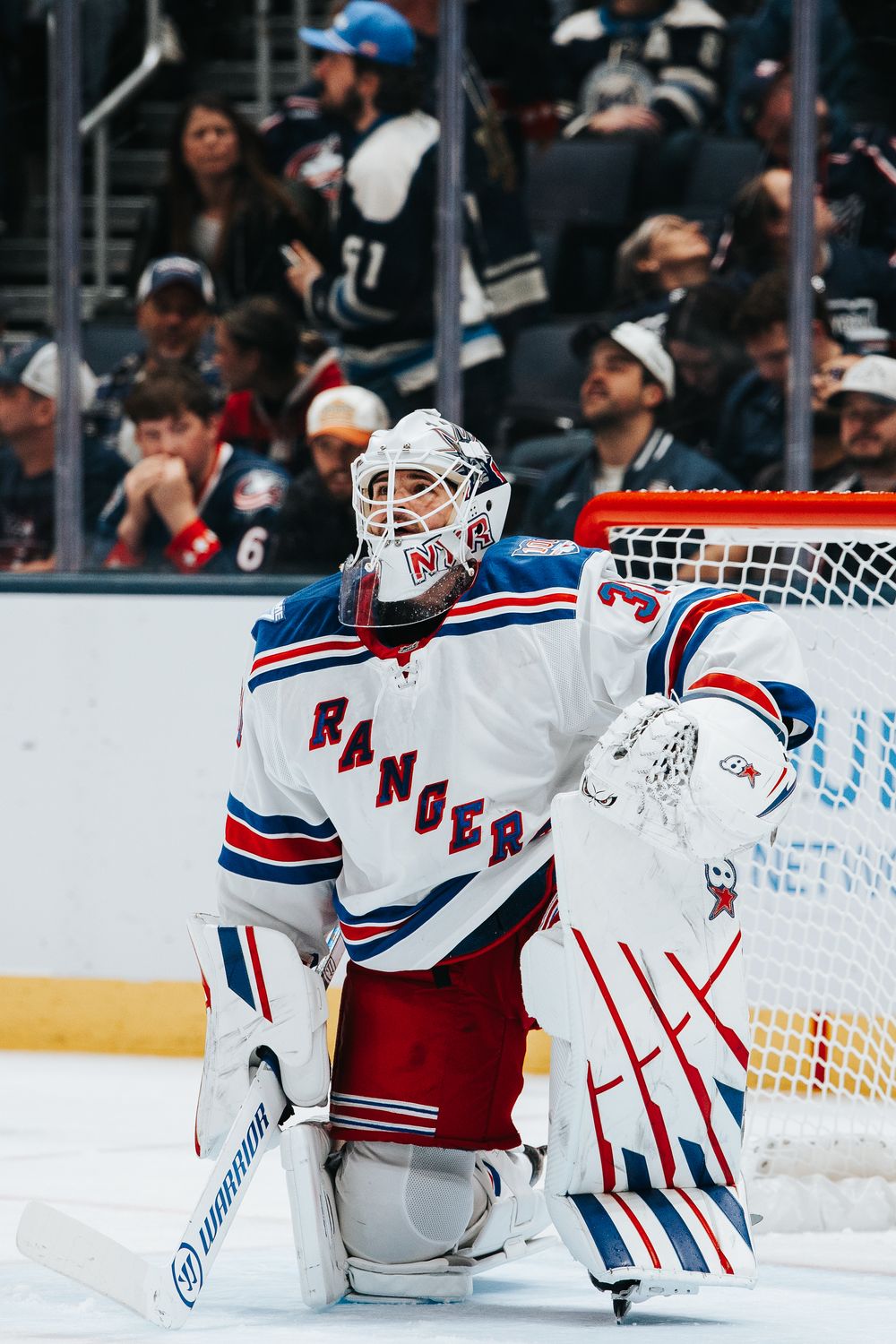 NHL goalie in New York Rangers white jersey and mask stands ready in goal during hockey game.