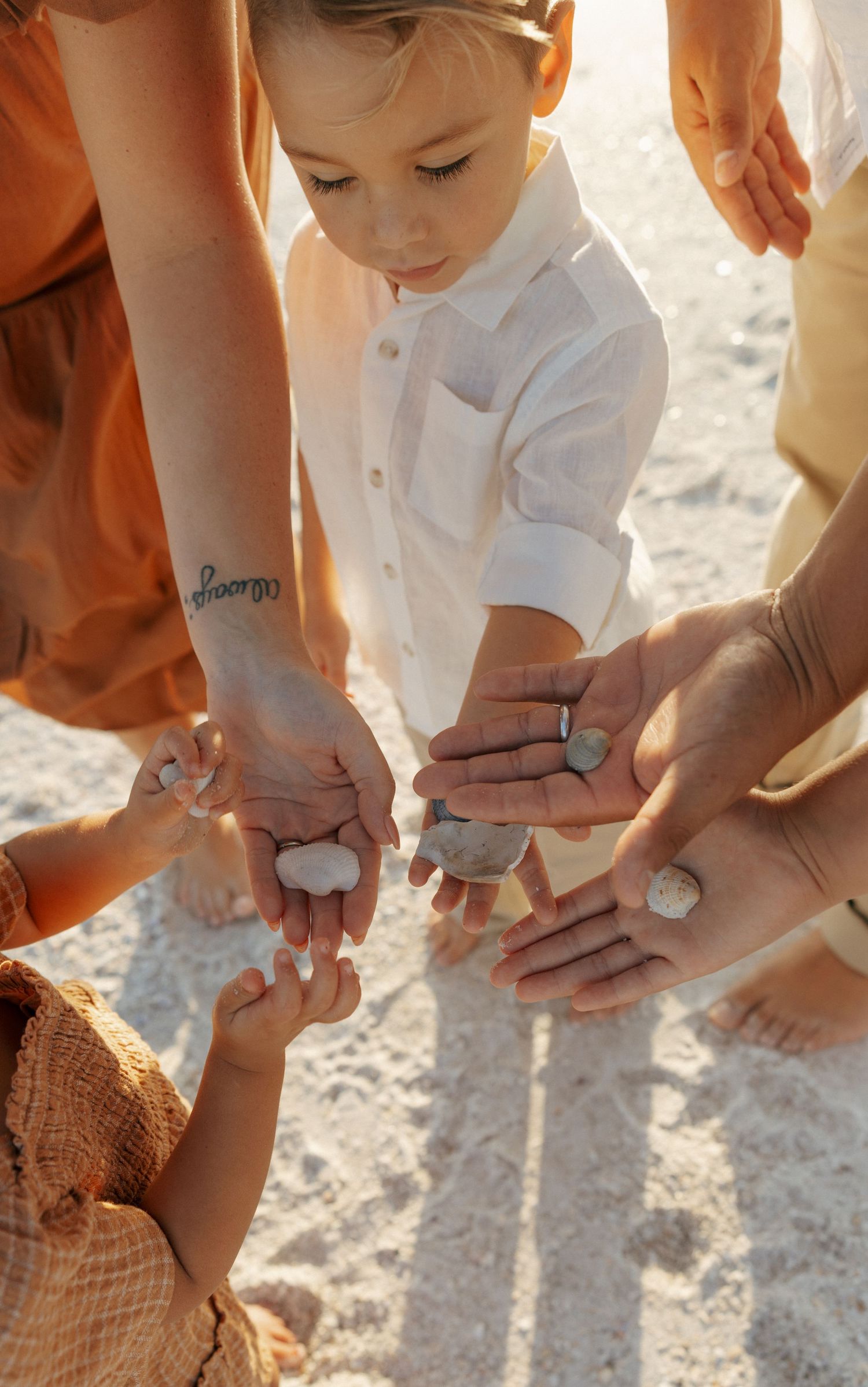 Lifestyle family photo of a young child standing on the beach surrounded by parents’ hands in warm natural light.