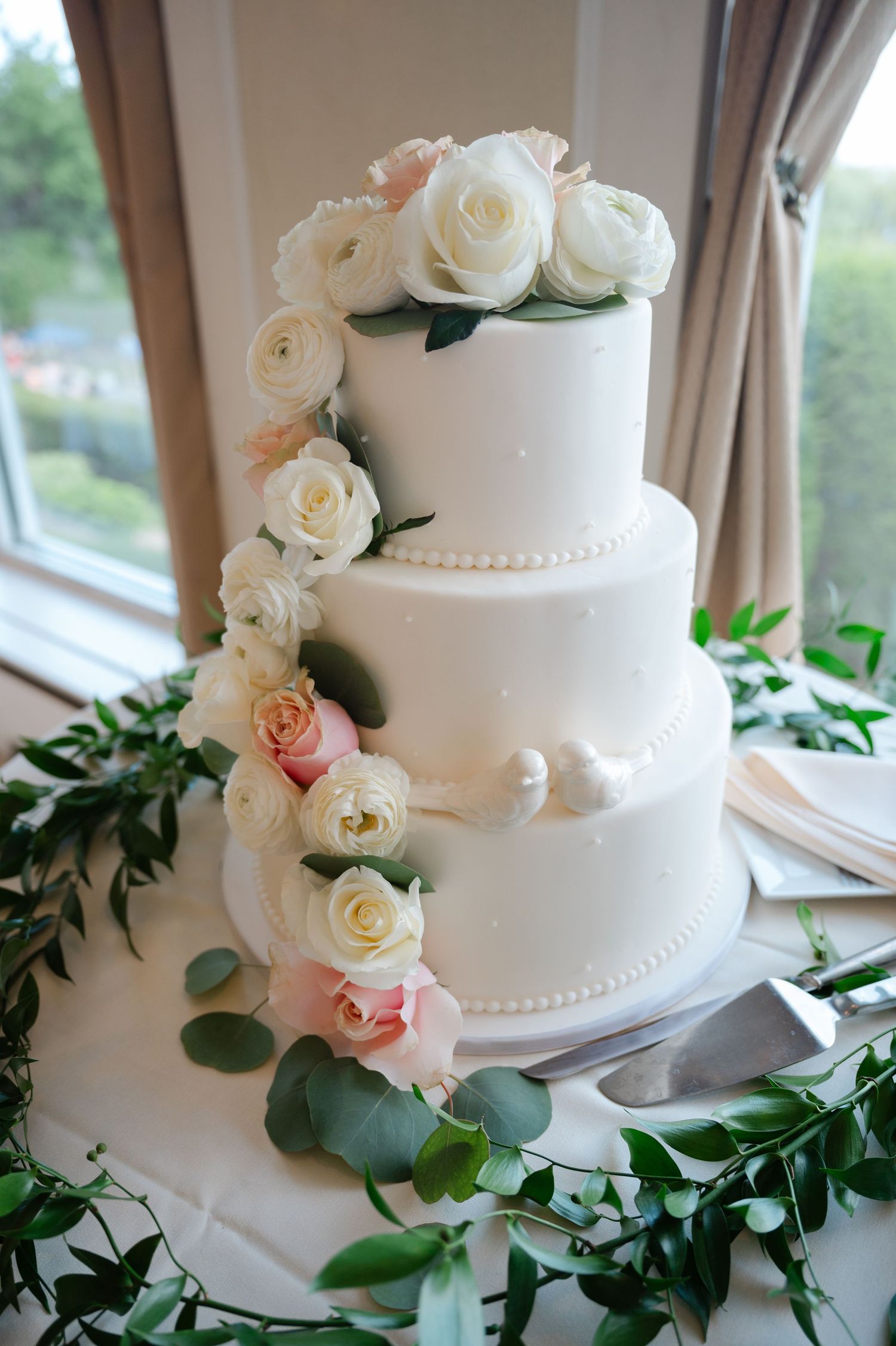 Three-tier white wedding cake adorned with pink and white roses, pearl details, and greenery garland on a white table.