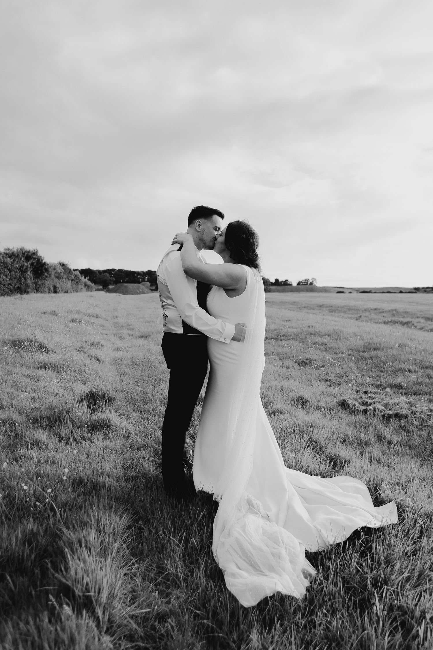 Black and white photo of a bride and groom embracing in a field, her gown flowing in the wind.
