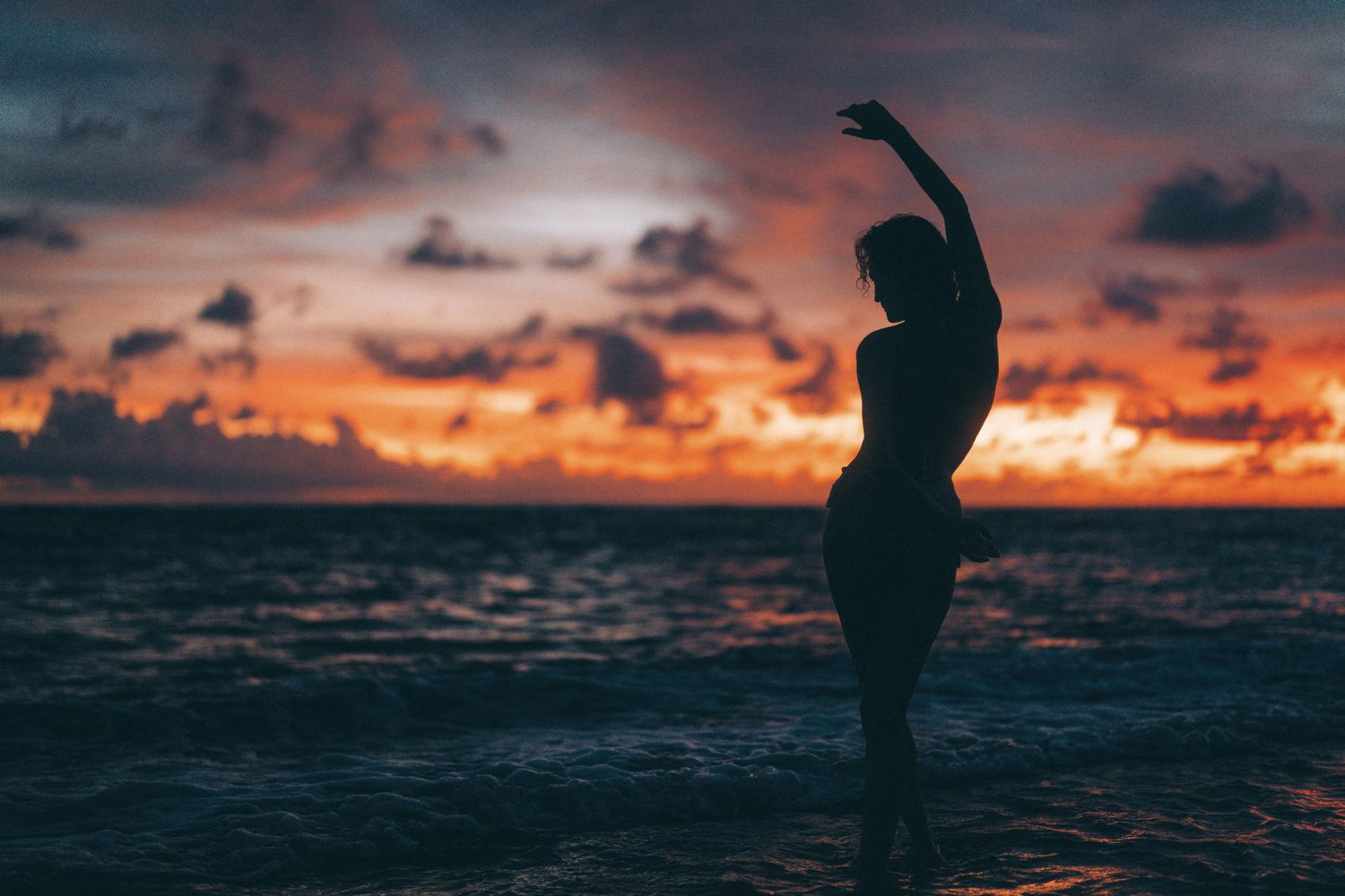 Silhouette of a woman dancing on a beach at sunset with vibrant orange and red clouds over the ocean.