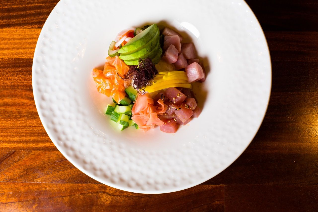 Elegant white plate with fresh tuna poke, avocado, salmon, and colorful garnishes on a wooden table.