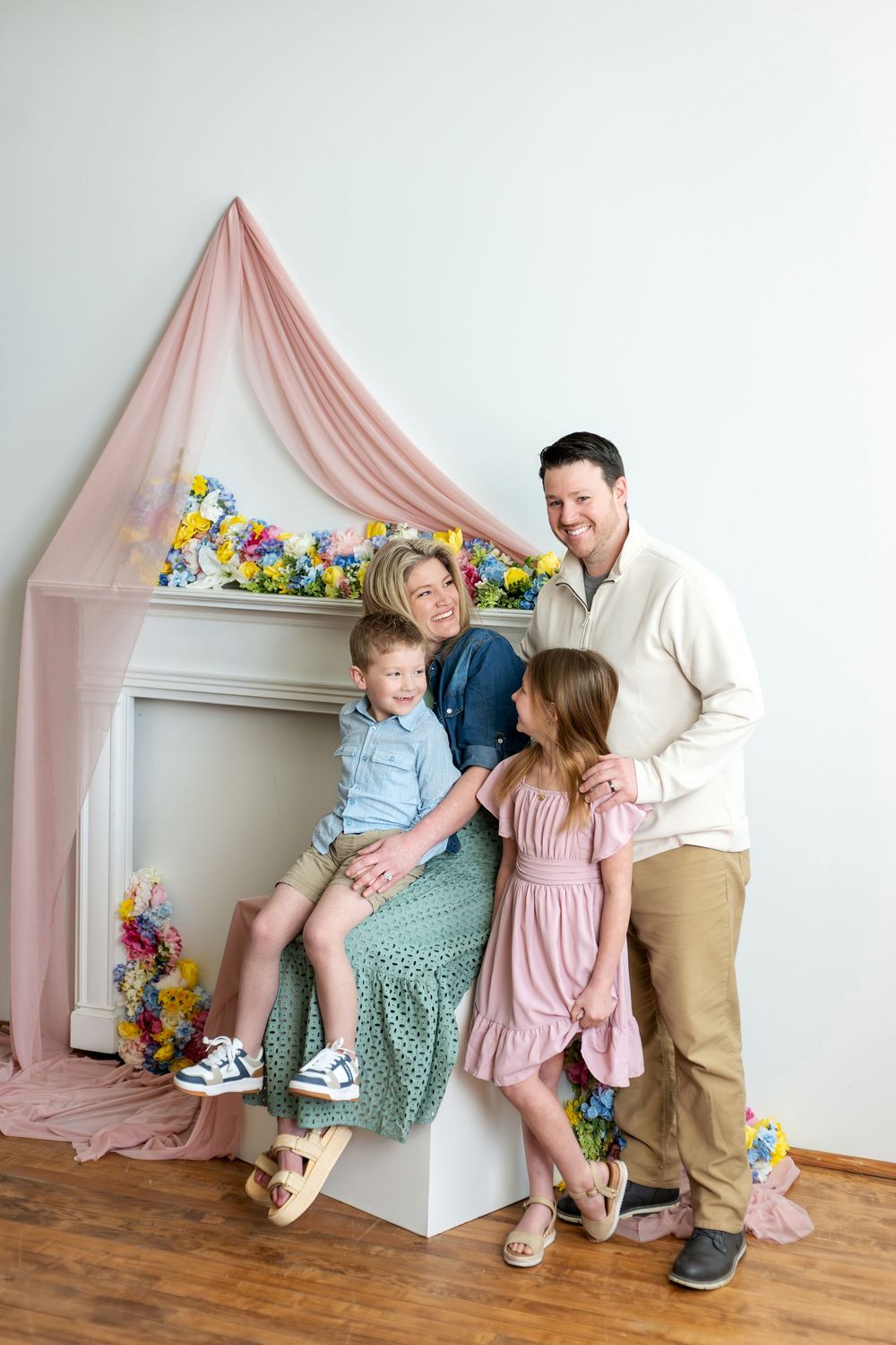 A happy family of four shares a candid moment in front of a spring-themed floral fireplace backdrop with pink draping.