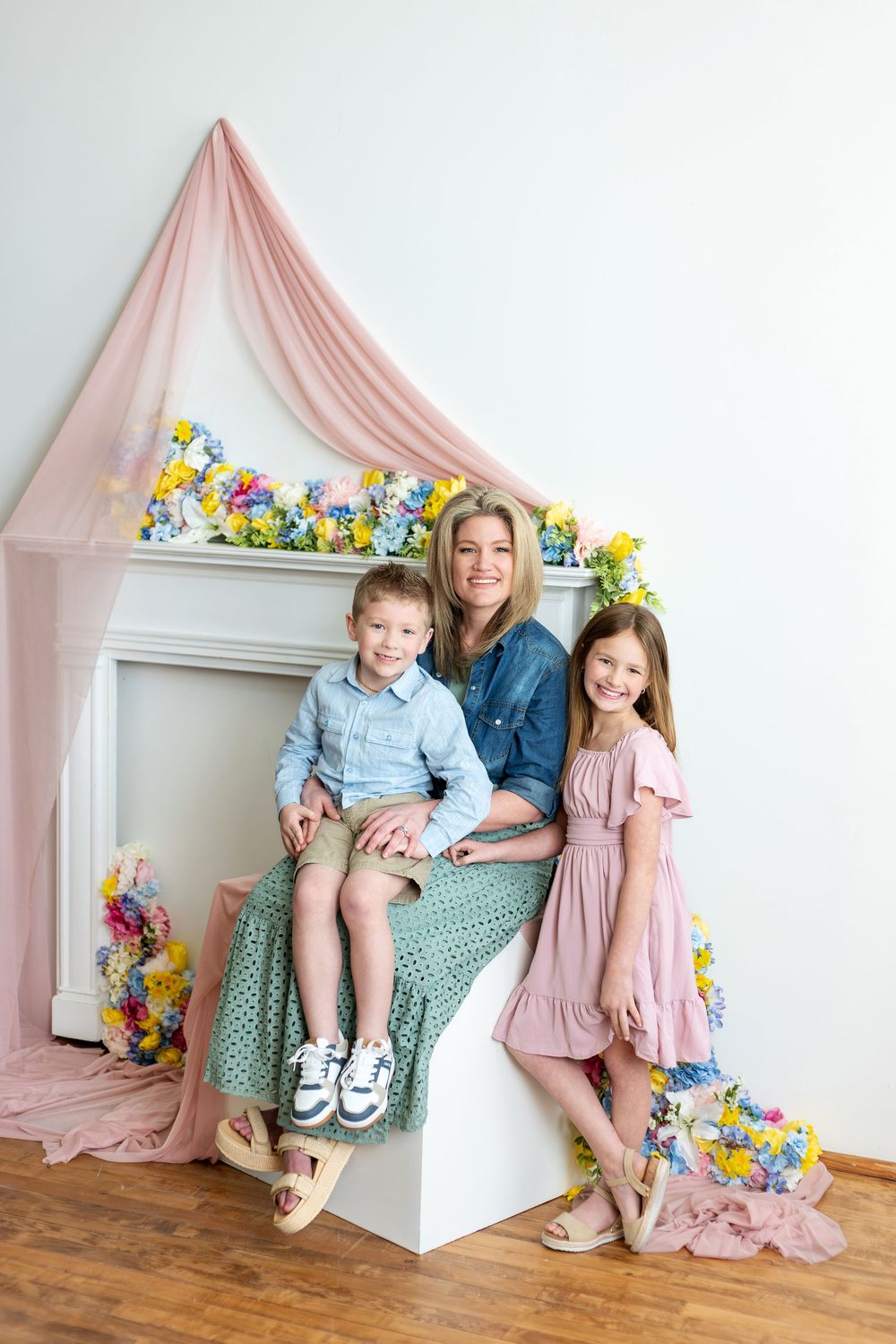 A mother sits with her two children in front of a white fireplace beautifully decorated with colorful spring flowers.