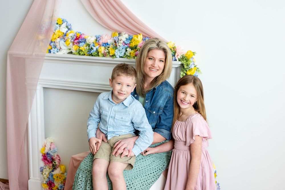 A smiling mother poses with her young son and daughter in front of a colorful spring floral fireplace decoration.