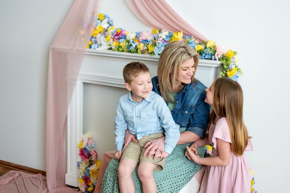Mother in denim shirt sits with young son and daughter near a colorful spring floral fireplace backdrop.