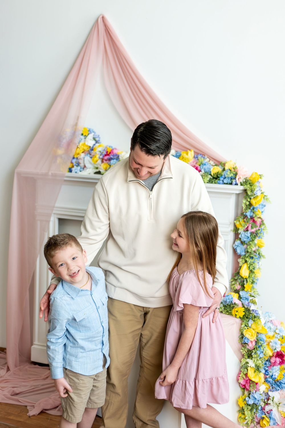 Father in cream sweater poses with two children before a floral spring backdrop with pink draping.