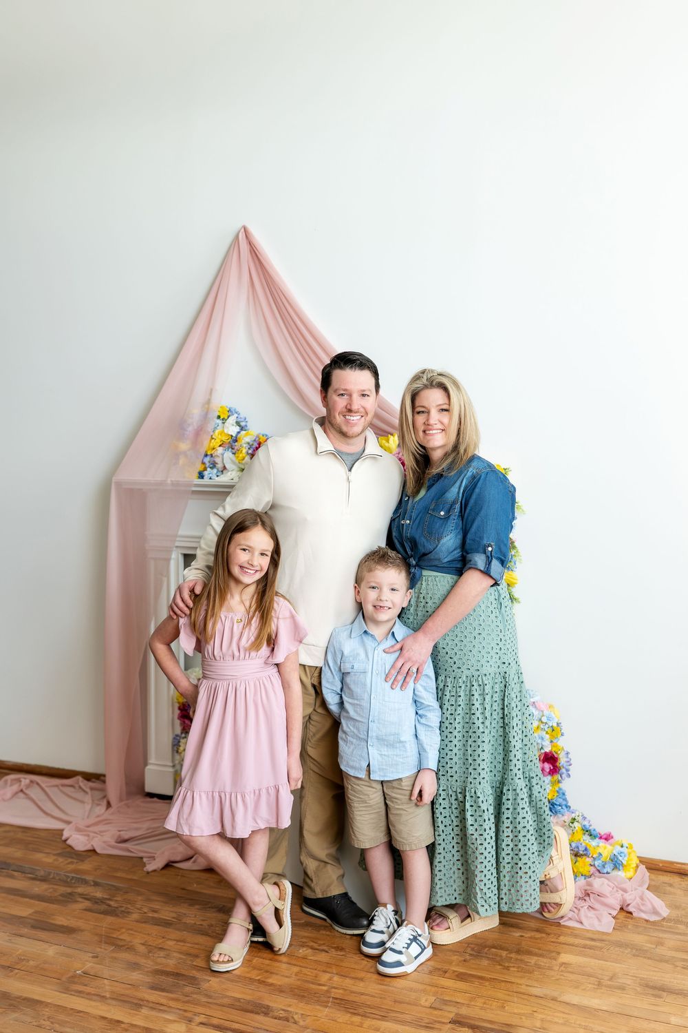 Happy family of four poses before a pink draped spring floral backdrop in coordinated pastel outfits.