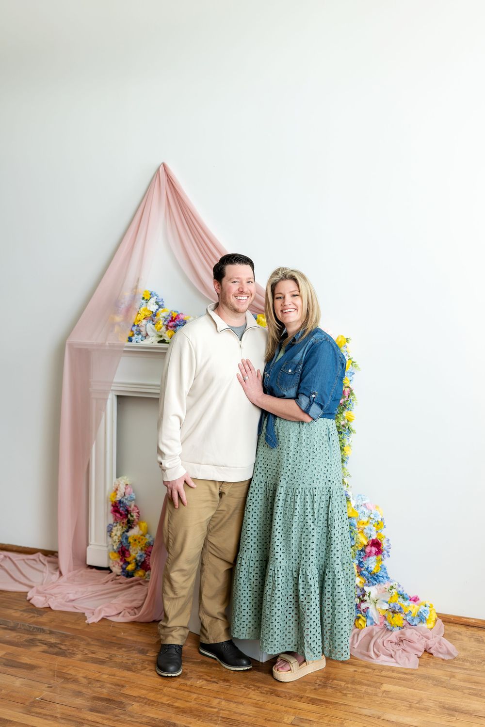Smiling couple stands together before a pink draped spring floral backdrop in a bright white studio.