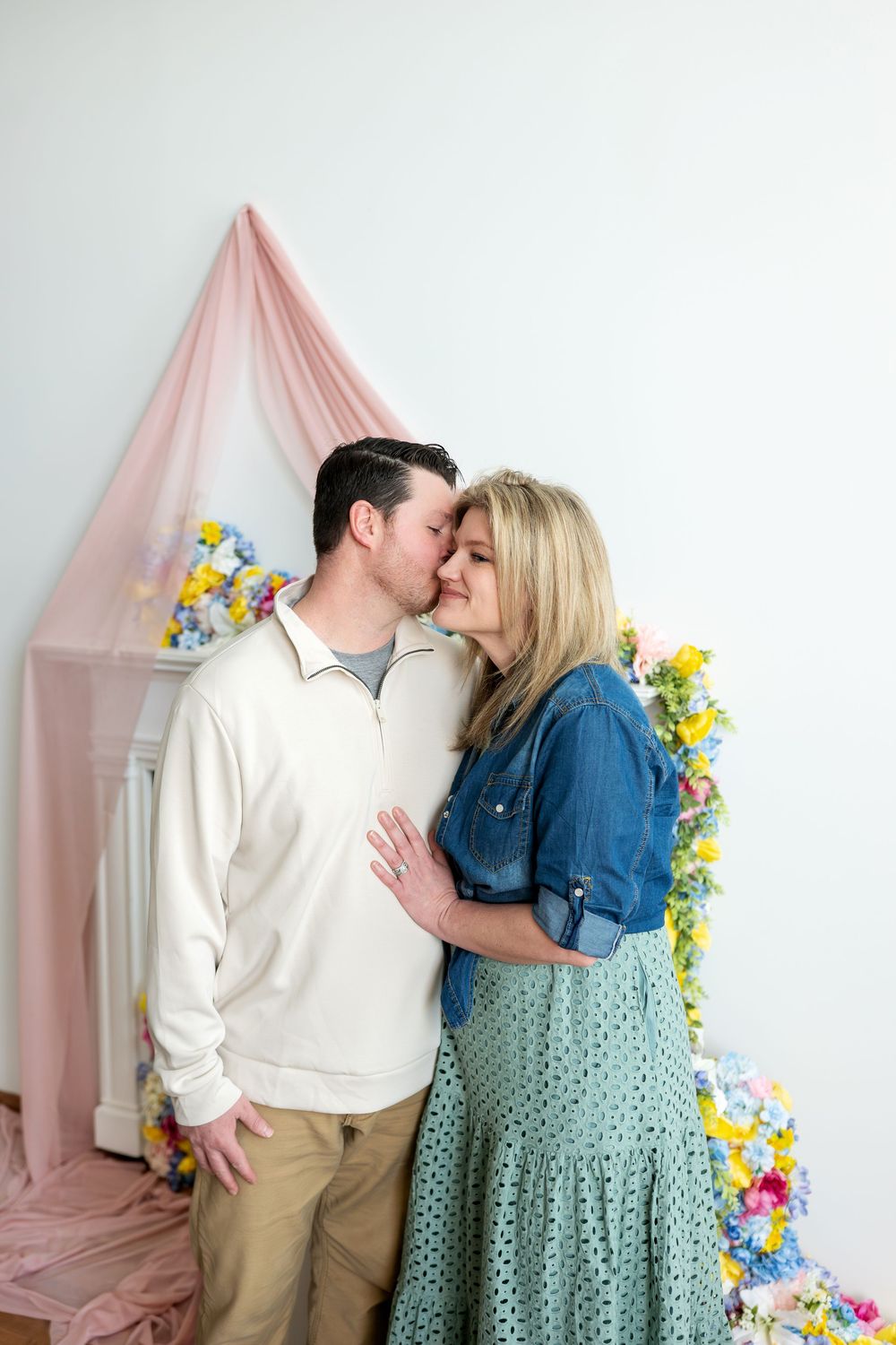 Couple sharing a kiss before a vibrant spring floral backdrop with pink draping in a photo studio.