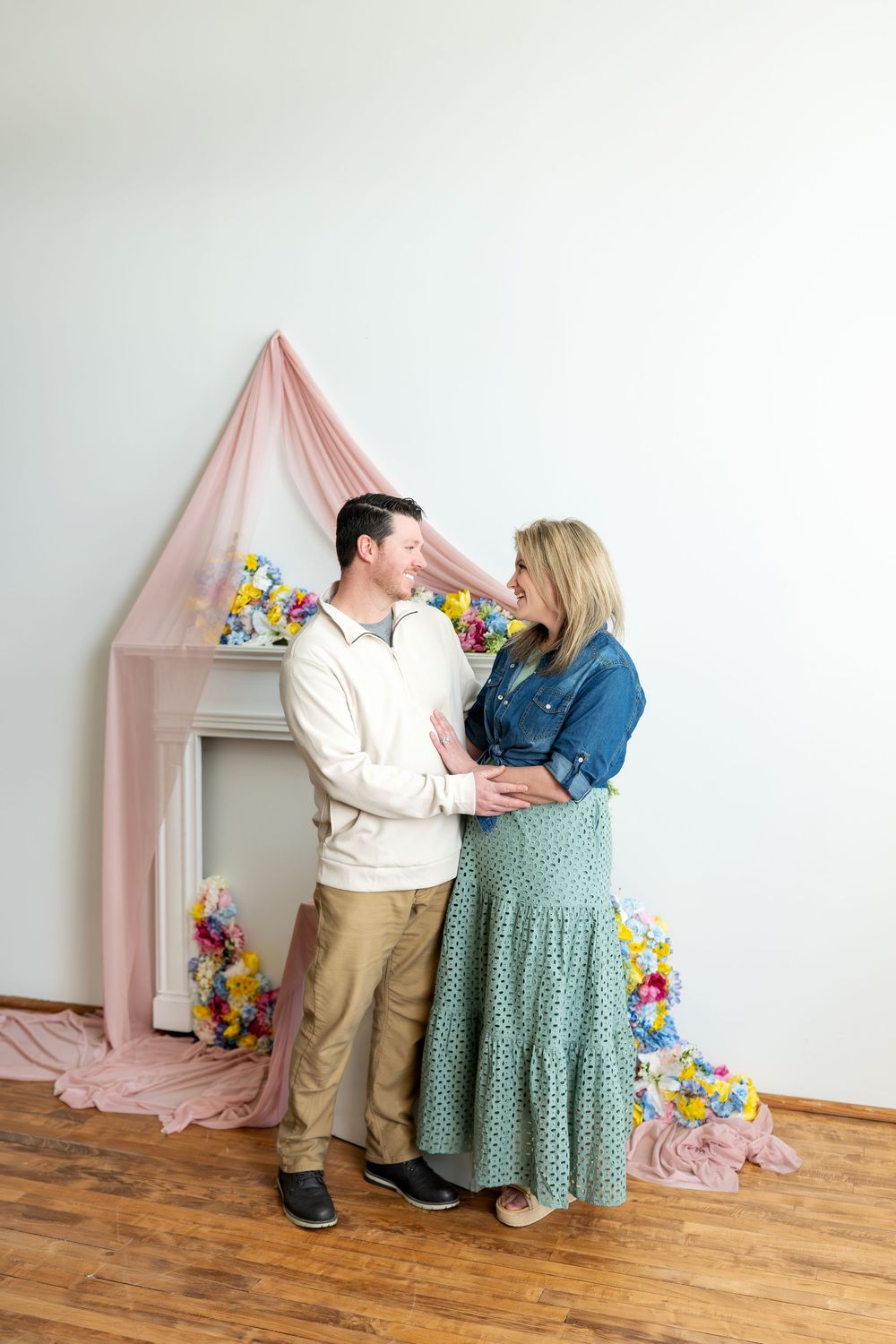 Couple smiling at each other before a pink draped spring floral fireplace backdrop in a bright studio.