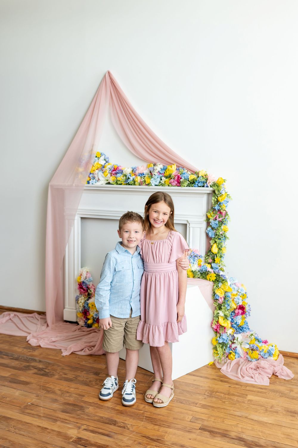 Young brother and sister in pastel outfits pose before a colorful spring floral fireplace backdrop.