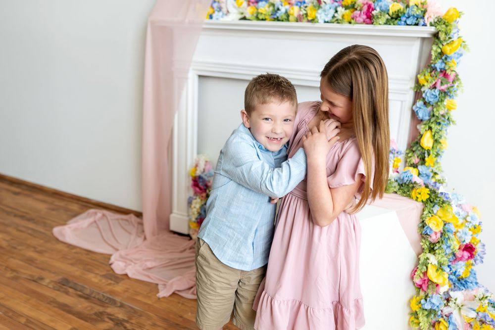 Two children hug and smile in front of a colorful flower-decorated white fireplace.
