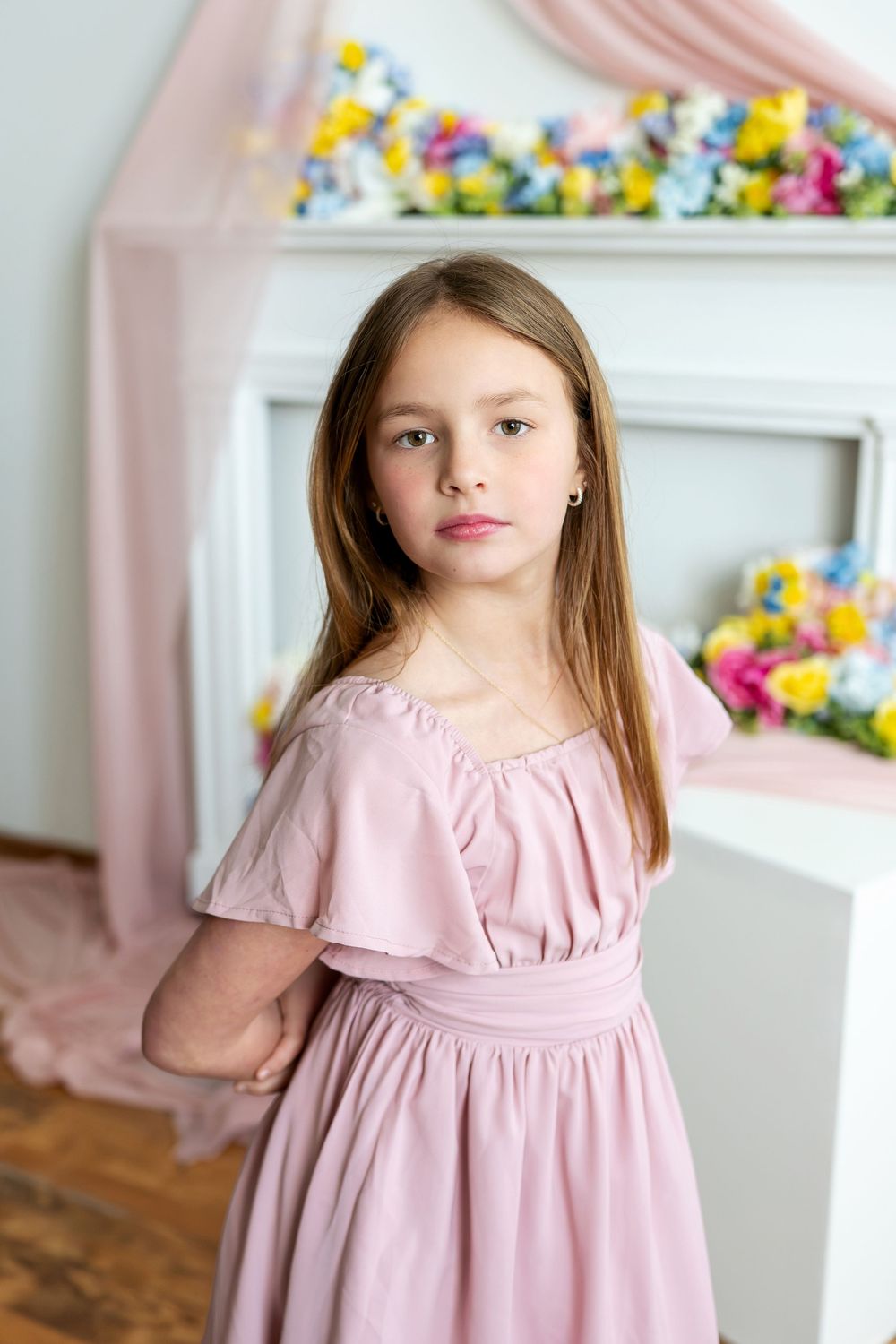 Young girl in pink dress stands pensively before a flower-adorned white fireplace backdrop.