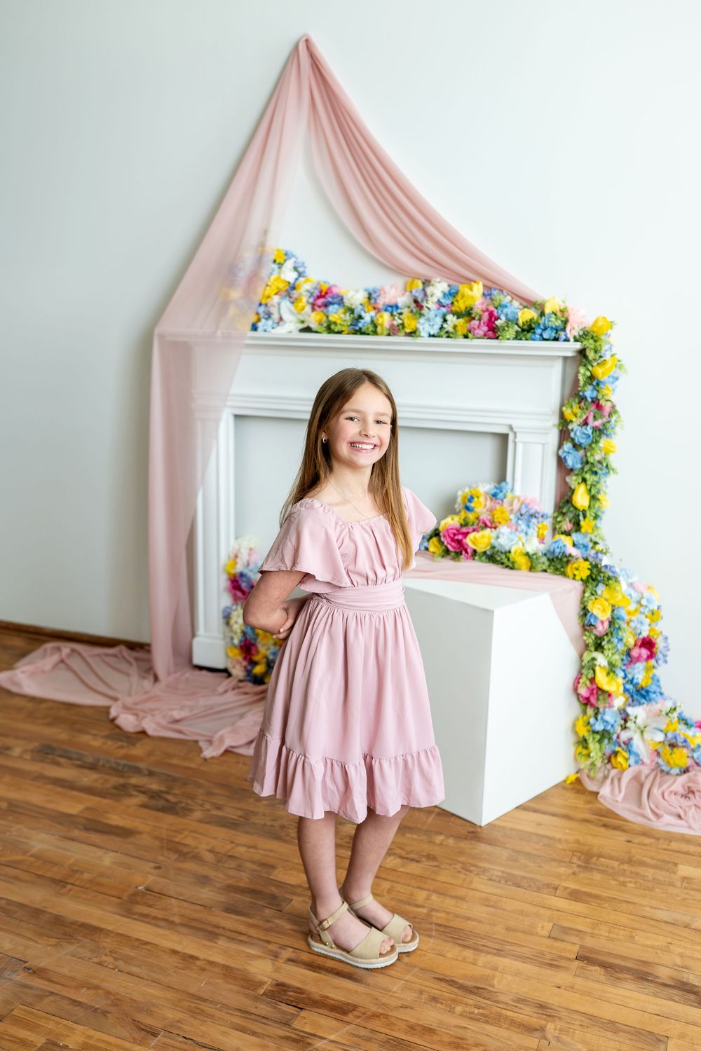 Happy girl in pink dress smiles broadly before a floral fireplace with pink canopy decor.