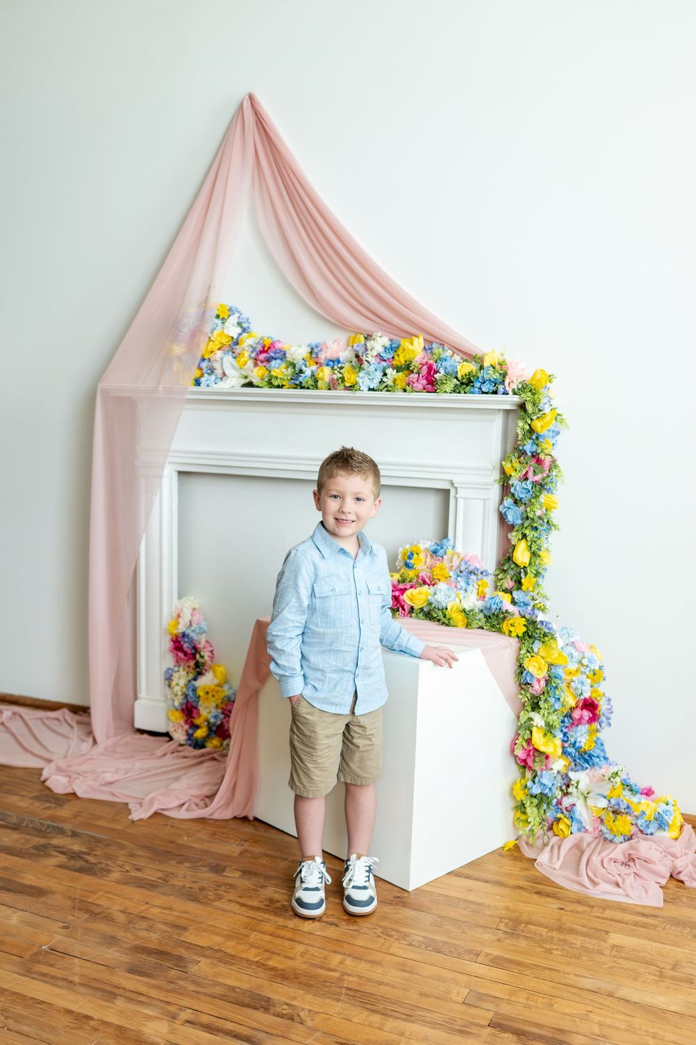 Smiling boy in blue shirt and khaki shorts stands by a flower-decorated white fireplace.