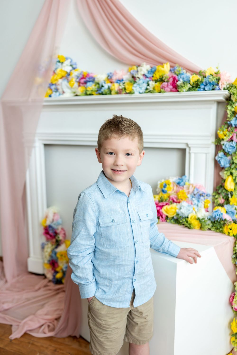 Young boy in light blue shirt smiles beside a colorful floral-decorated white fireplace.