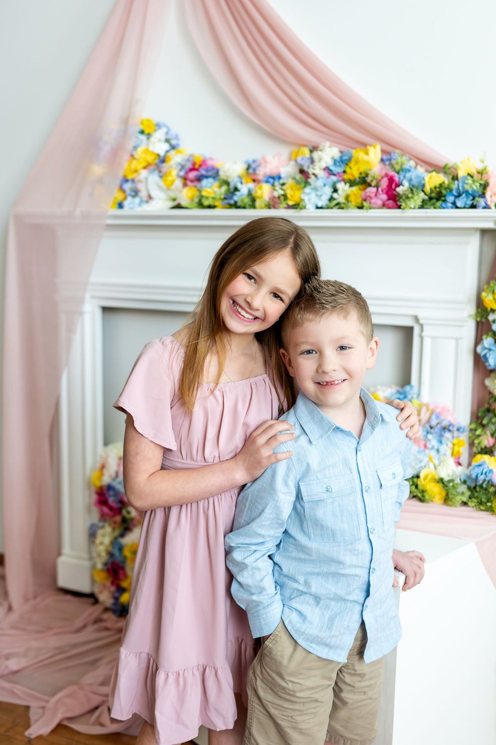 Smiling girl and boy pose together in front of a vibrant flower-decorated white fireplace.