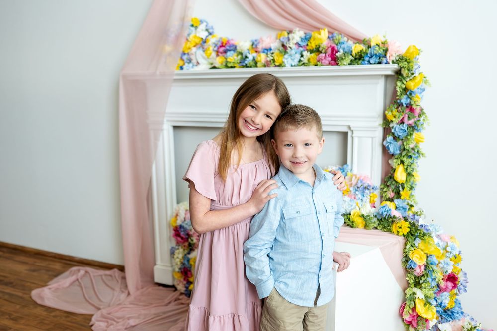 Sister and brother smile together before a colorful floral fireplace with pink draping.
