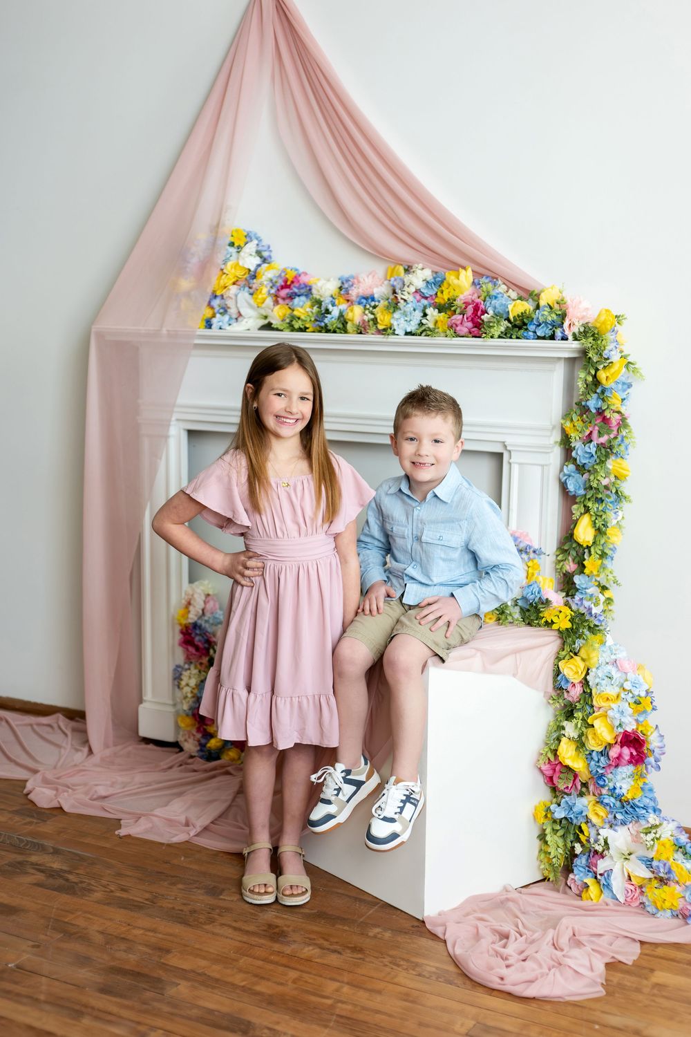 Smiling girl in pink dress and boy in blue shirt posing by colorful spring floral fireplace.