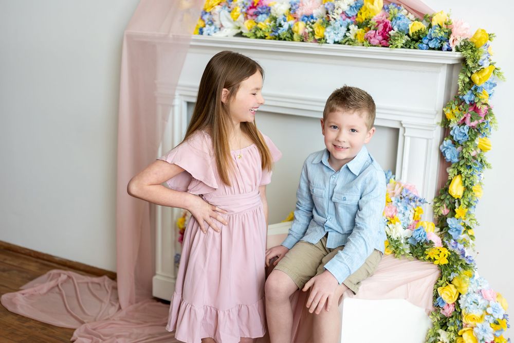 A girl in a pink dress and boy in a blue shirt stand together beside a white fireplace adorned with colorful spring flowers.