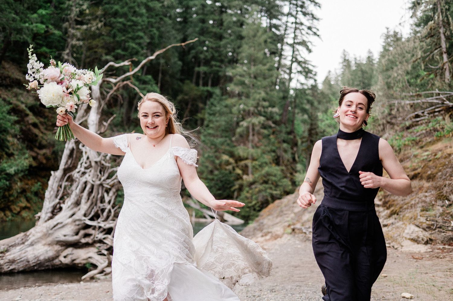 A couple in wedding attire shares a joyful moment together in a scenic forest setting with tall pine trees.
