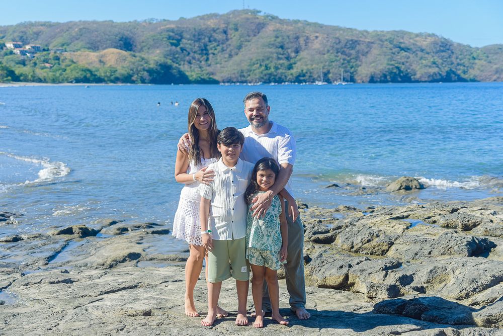 A group stands together on rocky coastal terrain with blue water and mountains in the background.
