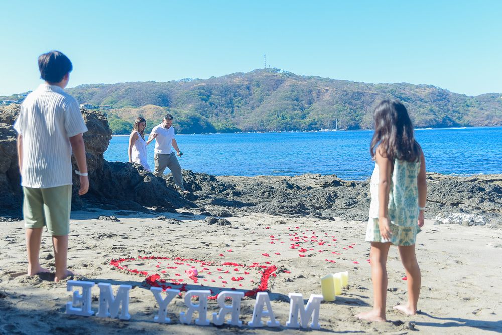 Beachside proposal setup with rose petals and letters arranged on rocky shore overlooking blue ocean and mountains.