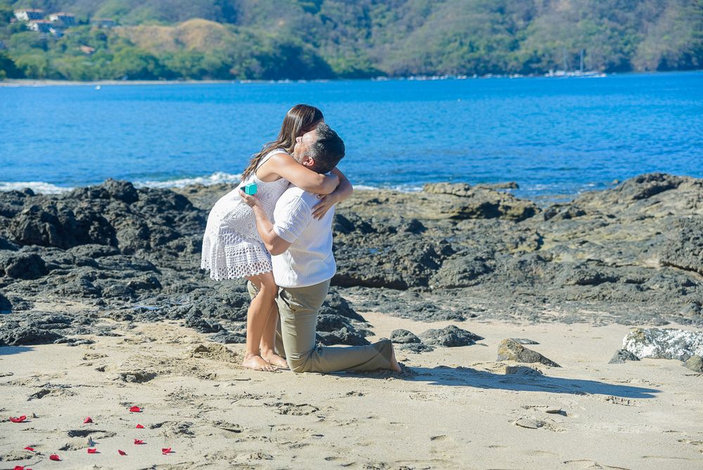 Couple enjoying a romantic moment on a scenic rocky beach with blue ocean waters and mountains in the background.