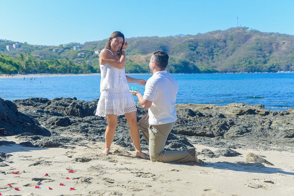 A romantic proposal scene on a rocky beach with blue ocean and mountains in the background.