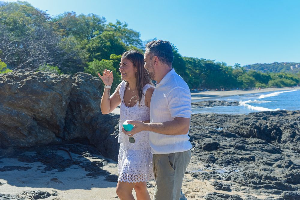 Couple enjoying a romantic moment together on a scenic rocky coastline with blue ocean and mountains in the background.