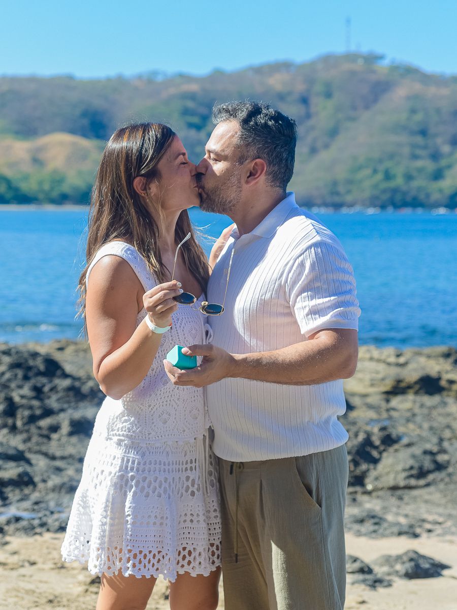 A romantic couple shares a tender moment by the ocean with mountains and blue water in the background.