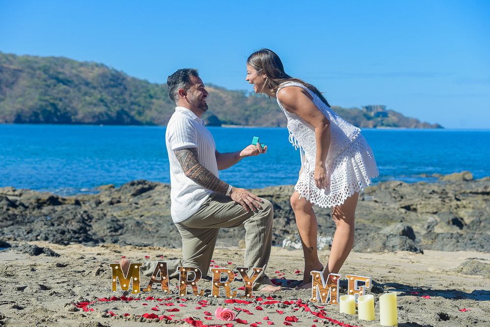A romantic proposal setup on rocky beach with candles and decorations against blue ocean backdrop.
