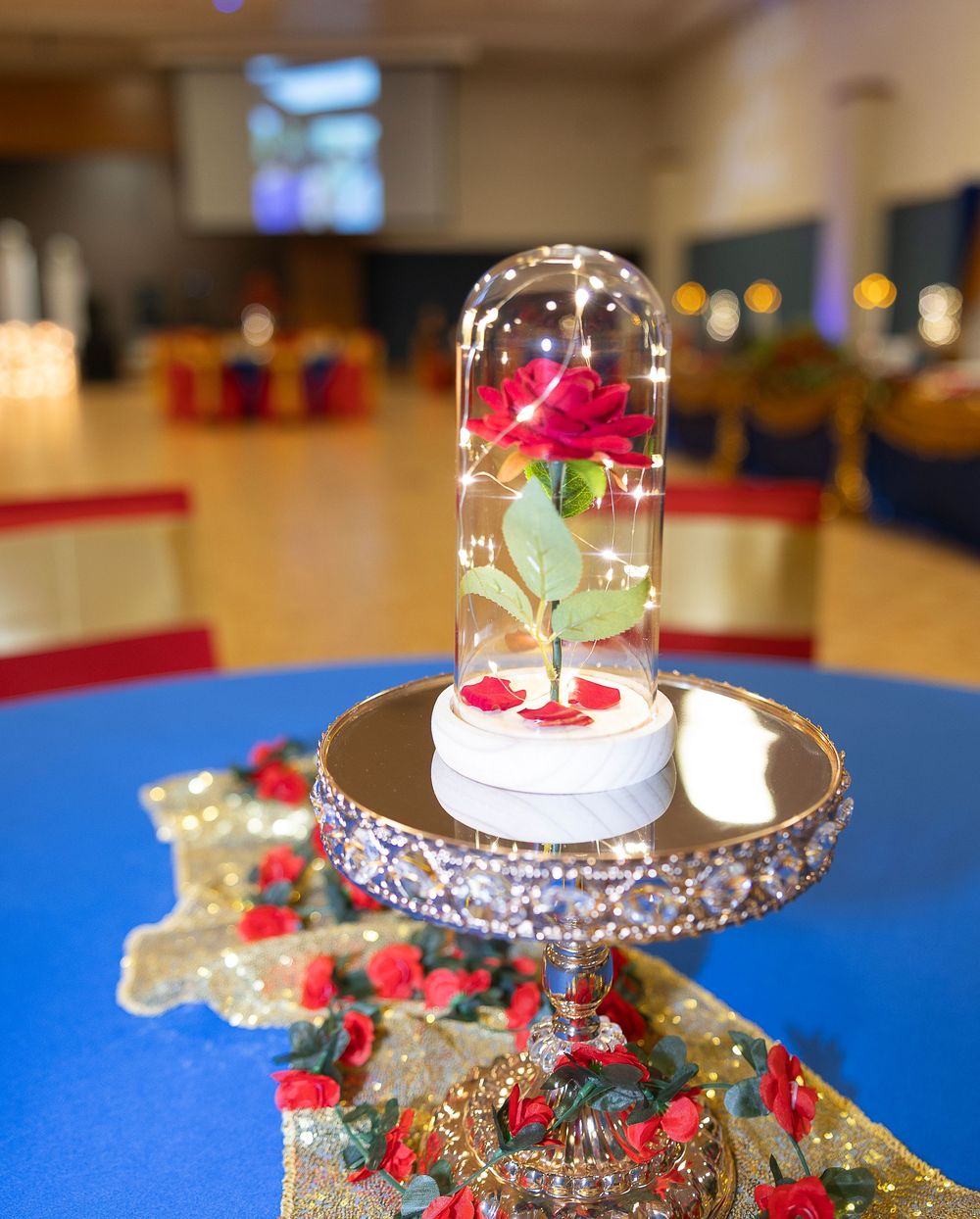 An elegant glass-domed display piece surrounded by red roses and greenery on a blue surface.